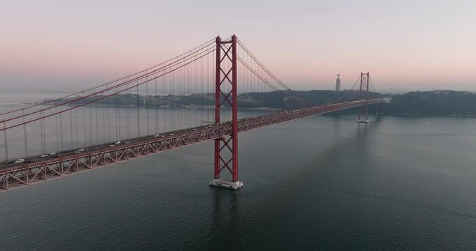 Aerial drone shot towards the 25th April Bridge, Ponte 25 de Abril, and Cristo Rei in Lisbon, Portugal, Europe. Dusk after sunset with pink and orange sky. Shot in ProRes 422 HQ