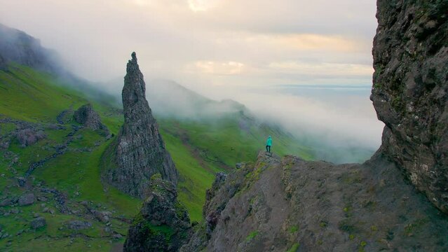 AERIAL: Woman climbed to the viewpoint where she admires the misty Cathedral Rock. Young adventurer is exploring unique natural attractions with picturesque dramatic landscape on the Isle of Skye.
