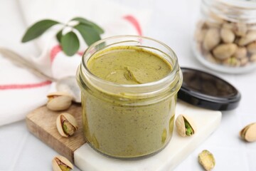 Tasty pistachio cream in jar and nuts on white table, closeup