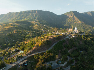 An aerial view of a mountainous landscape at sunrise, showcasing a serene temple complex with a prominent white Buddha statue, nestled among the lush greenery.