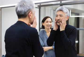 鏡を見ながら顔を触っているミドル男性/女性がスキンケアを教えている/メンズビューティー