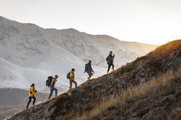 Group of young active hikers with backpacks walks uphill in mountains at sunset