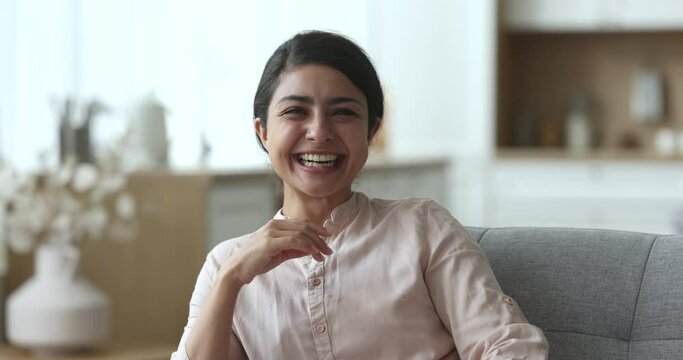 Beautiful young Indian woman posing for head shot portrait at home, looking at camera, getting happy, cheerful, laughing with hand at chin, sitting on sofa at home, enjoying leisure, comfort