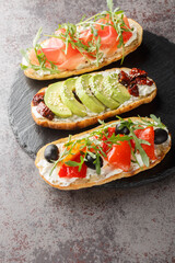 set of bruschetta sandwiches with arugula, ham, sun-dried tomatoes, avocado, cream cheese and roasted peppers close-up on a slate board on the table. Vertical