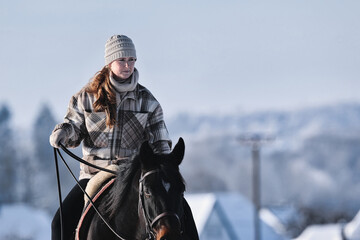 A young red-haired woman rides her horse at a walk across a Schebeckte meadow with a snowy village...