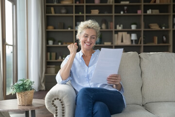 Happy elderly woman sit on couch in living room, hold papers, receive bank notice about loan...