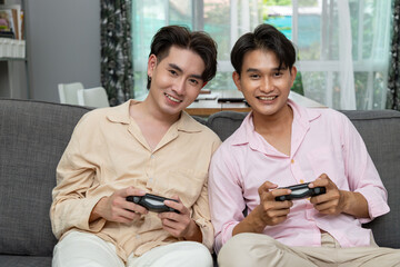 Cute happy smiling Asian LGBT homosexual men or gay couple sitting on sofa, holding joypad, playing game together.