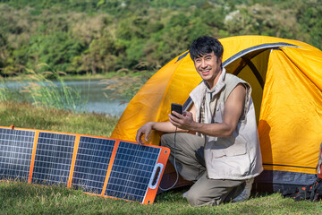 Young Asian male man sitting in front of camping tent beside lake and mountain with solar cell, charging mobile phone. Looking at camera.