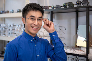 Portrait of Young Asian male man choosing glasses in optical shop, trying eyeglasses. Eyecare concept. Looking at camera.