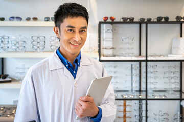 Portrait of young Asian male man optician in optical shop store, holding digital tablet. Eyecare and shopkeeper concept. Looking at camera.