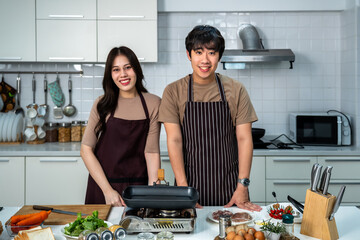 Portrait of young Asian couple male man female woman cheerful standing and preparing food for meal, enjoy cooking, in kitchen at home. Looking at camera.