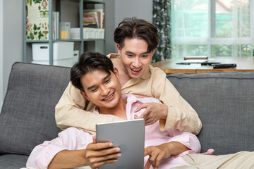 Cute happy smiling Asian LGBT homosexual men or gay couple sitting and reclining on sofa, embracing and using tablet together.