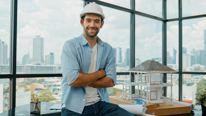 Portrait of architect engineer in casual outfit smile at camera while crossing arms. Engineer looking at camera and sitting with arms folded near house model while wearing safety helmet. Tracery - obrazy, fototapety, plakaty