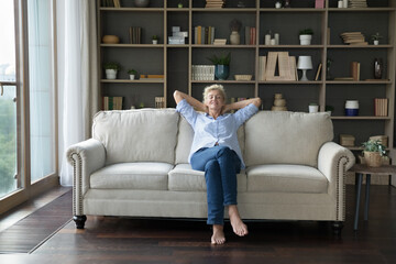 Mature female put hands behind head relaxing on comfortable fashionable sofa in spacious living...