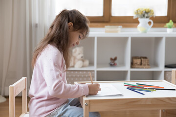 Little focused girl sit at table drawing pictures looks engrossed in painting process in...