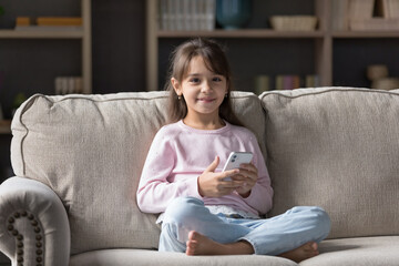 Little girl sit on couch in living room with modern smartphone, smile, look at camera, play on-line...