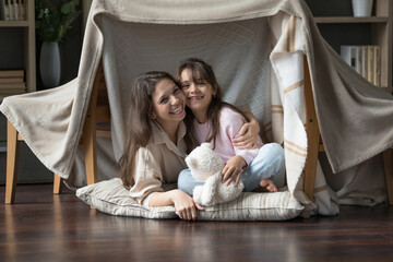 Fototapeta na wymiar Happy young woman sits on soft cushion under handmade teepee with cute little daughter play toys spend free time together in cozy, modern flat. Family ties, motherhood and playtime with kids at home