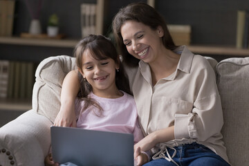 Laughing mom and kid girl watching on-line movie using laptop and streaming subscription services,...