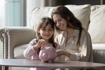 Beautiful little 6s daughter put coins into pink piggybank, smile spend time at home with caring...