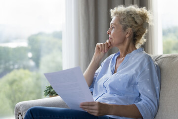Thoughtful mature woman sitting on sofa at home, hold papers looking away, think of financial...