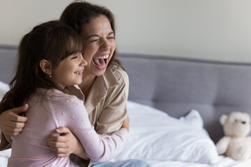 Overjoyed mother hugging laughing little 5s daughter sit on bed in bedroom, look away, having fun in morning at home, enjoy playtime, have playful mood feel happy spend family leisure indoor, close up
