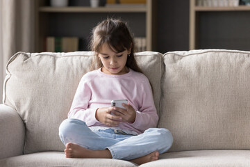 Little 6s engrossed Latina girl rest on sofa with smart phone spend free time on internet, watching...