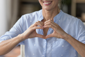Close up crop view of smiling mature woman joining fingers makes symbol of love, showing heart...