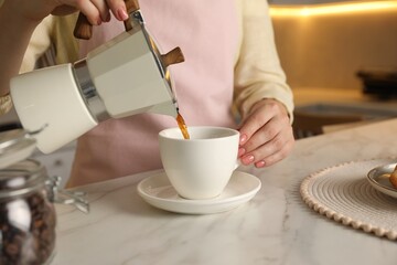 Woman pouring aromatic coffee from moka pot into cup at white marble table, closeup