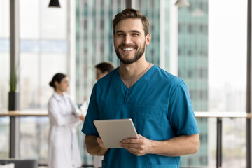 Cheerful handsome surgeon doctor man in blue uniform holding digital tablet computer, looking at camera, smiling, posing for portrait in clinic hall, promoting modern technology in medical job - obrazy, fototapety, plakaty