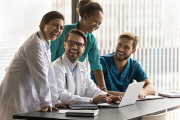 Group of cheerful diverse doctors laughing at laptop, relaxing on job meeting, testing medical application for work, talking, having fun, brainstorming, enjoying online communication at workplace