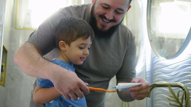 Father putting tootpaste on a toothbrush, helping and teaching his little son to brush his teeth in the bathroom in the morning