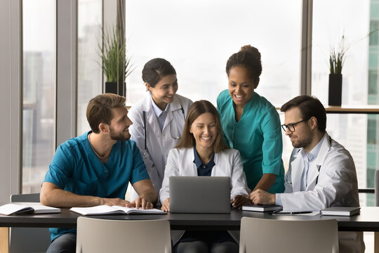 Happy successful young multiethnic doctors meeting at laptop, talking on video call to colleagues, smiling, laughing, standing at medical team leader using computer