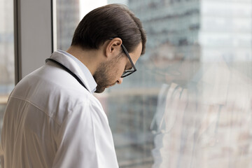 Tired frustrated young doctor man standing at window with closed eyes and head bended down,...