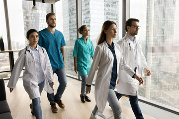 Diverse young doctors in white and blue uniforms walking past camera in city clinic office hall,...