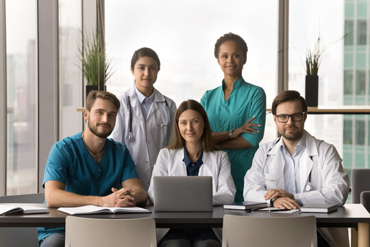 Positive diverse team of attractive young practitioners, surgeons, nurses posing together in hospital office, sitting and standing at table with laptop, looking at camera for portrait