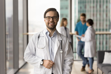 Happy successful medical practitioner man in uniform and glasses posing with team meeting behind, looking at camera, smiling, laughing, promoting doctor occupation, healthcare checkup