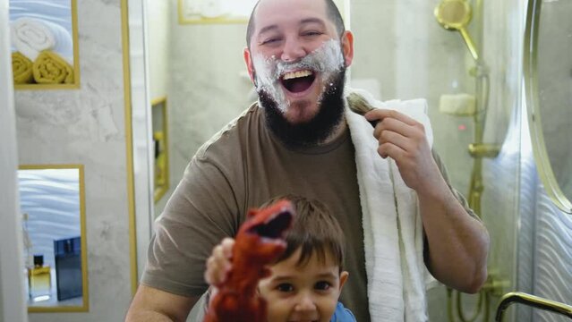 Portrait of a happy smiling dad shaving looking at the camera while his toddler plays with a toy dinosaur