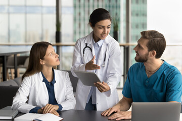 Beautiful young Indian doctor woman holding tablet, talking with colleagues, discussing diagnosis, giving consultation of expert. Multiethnic practitioners using gadgets for work discussion