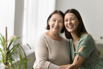 Naklejka premium Cheerful elder mature mother and beautiful adult daughter woman hugging with heads touch, sitting close, looking away with toothy smiles, posing for shooting, enjoying family meeting