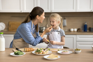 Cheerful mom feeding cute preschool kid girl while cooking in kitchen, giving slice of cheese kid...