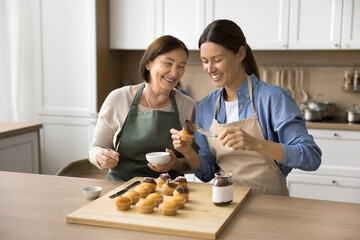 Cheerful senior mom and adult daughter woman in aprons enjoying baking activity in home kitchen,...