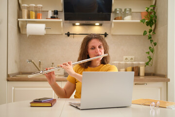 A woman with a laptop playing a flute is sitting at a table in a home kitchen. An adult female businesswoman works from home, a remote office