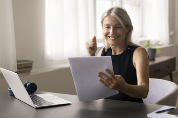 Happy attractive middle aged woman sit at table read postal letter correspondence, get good news in...
