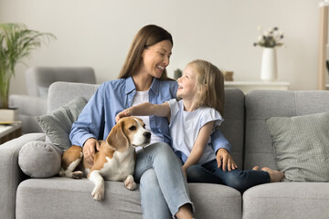 Happy beautiful mother embracing kid and touching dog on home sofa, talking to little daughter girl, smiling, laughing, relaxing in cozy home interior, spending family leisure with kid and pet