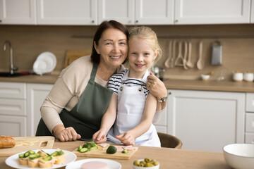 Happy mature grandma and little grandkid girl in aprons preparing sandwiches, cooking fast food in...