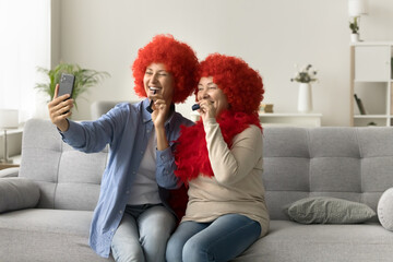 Cheerful senior mother and adult daughter woman in funny red clown wigs using smartphone for selfie, video call communication, celebrating birthday at home, blowing into party horn, laughing