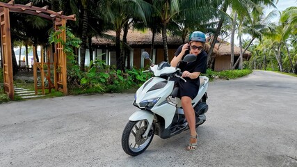 A person in a blue helmet sits on a white scooter, talking on the phone, with a tropical background suggesting a vacation or leisure concept