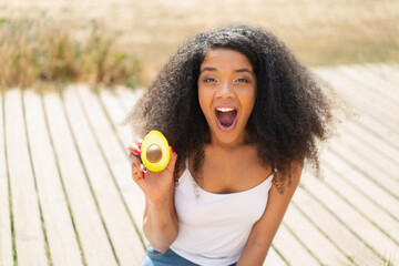 Young African American woman holding an avocado at outdoors with surprise and shocked facial...