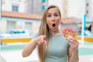 Young blonde woman taking a lot of money at outdoors surprised and pointing front