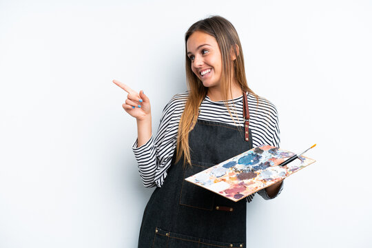 Young artist caucasian woman holding a palette isolated on white background pointing finger to the side and presenting a product